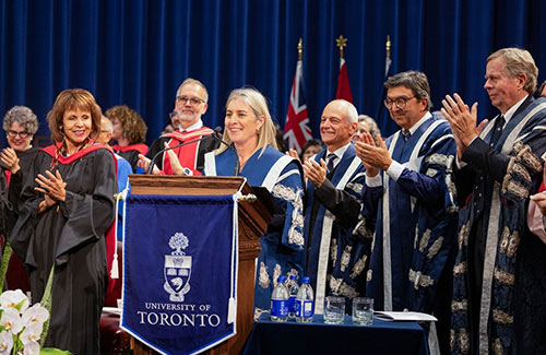 President Melanie Woodin at a podium, surrounded by U of T staff in formal scholar gowns