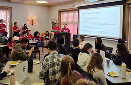 A group of students in red shirt stand in front of a lectern with a screen behind them while students eating at tables watch.