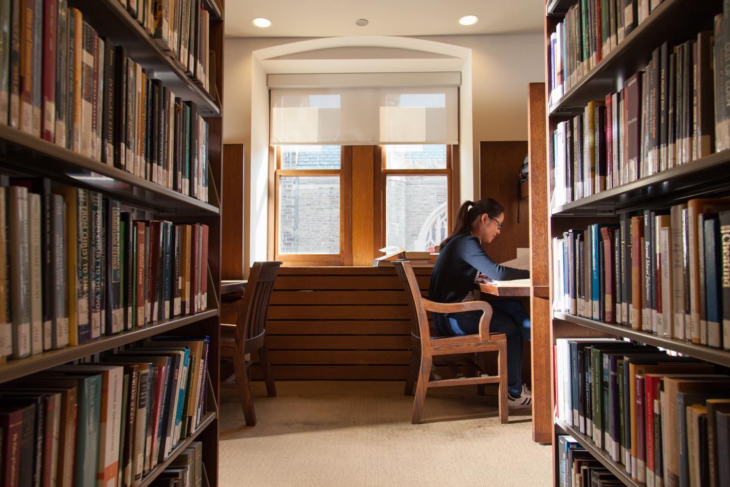 Student studying in a carrel in the sunlit John W. Graham Library with a large window in the back ground and shelves filled with books to the left and right in the foreground.