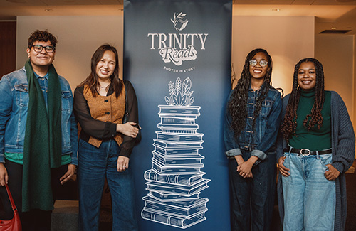 Four people stand on either side of a tall banner that says "Trinity Reads" with illustration of books