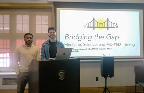 Two male students stand at a lectern beside a screen with the presentation title "Bridging the Gap: Medicine, Sciences and MD-PhD Training"